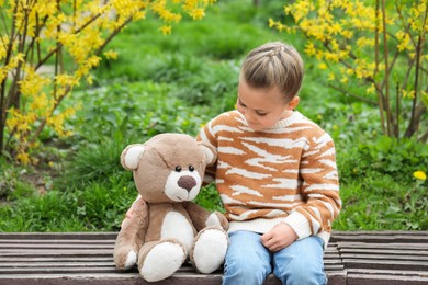 Little girl with teddy bear on wooden bench outdoors