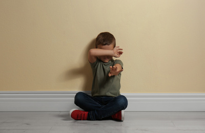 Photo of Scared little boy closing face with hand on floor near yellow wall. Child in danger