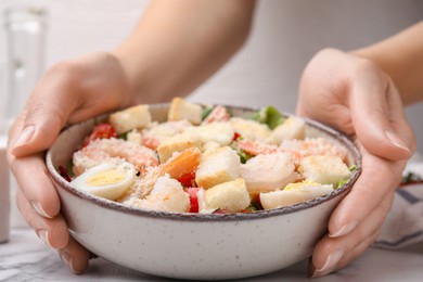 Woman holding bowl of delicious Caesar salad with shrimps at white table, closeup