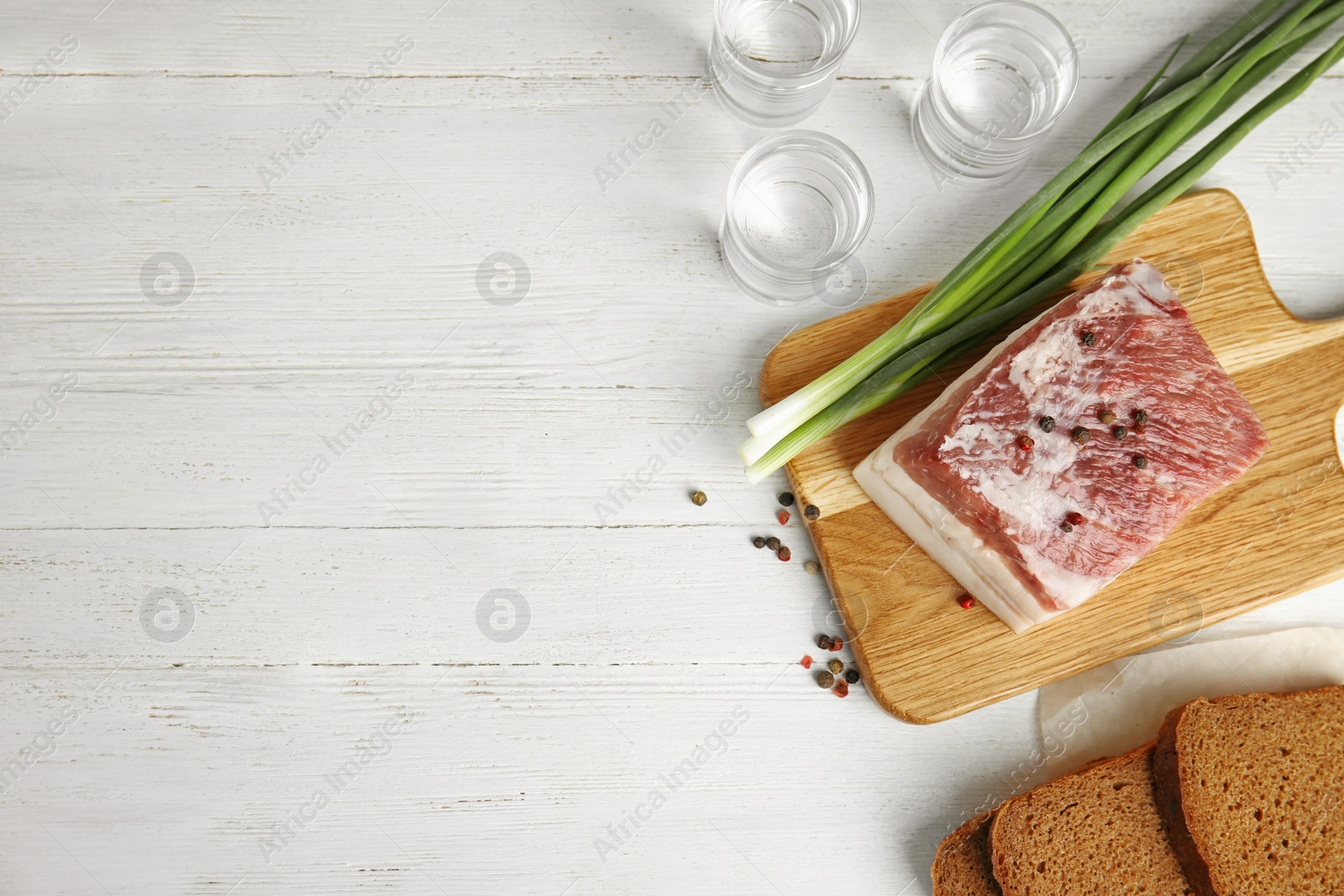 Photo of Board with pork fatback, onion, bread and shots of vodka on white wooden background, flat lay. Space for text