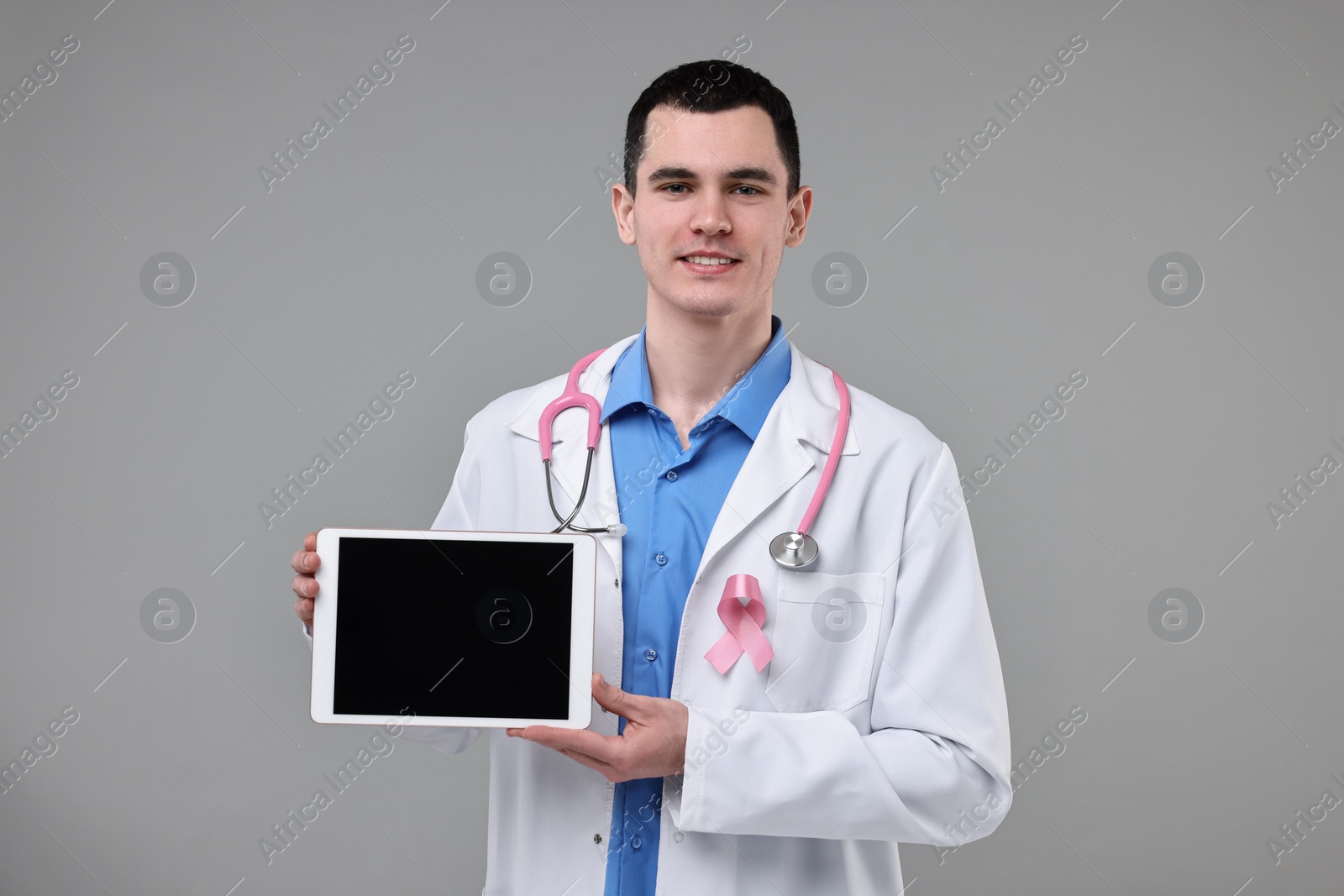 Photo of Smiling mammologist with pink ribbon and tablet on grey background. Breast cancer awareness