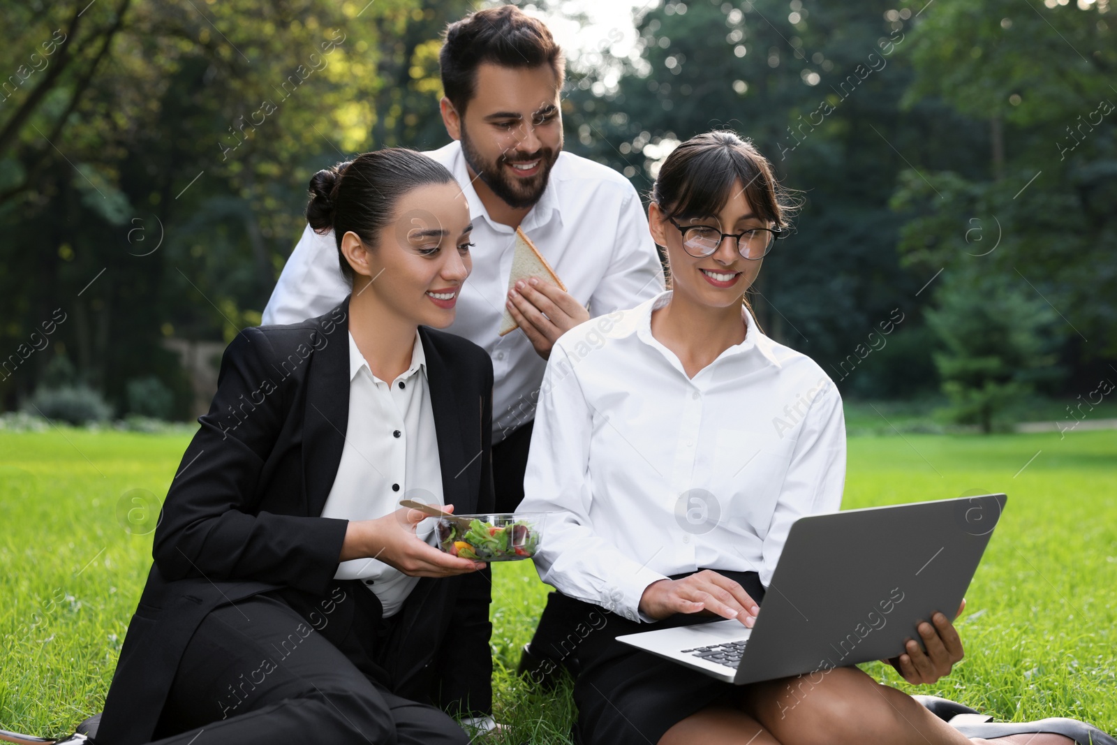 Photo of Happy colleagues with laptop having business lunch on green grass in park