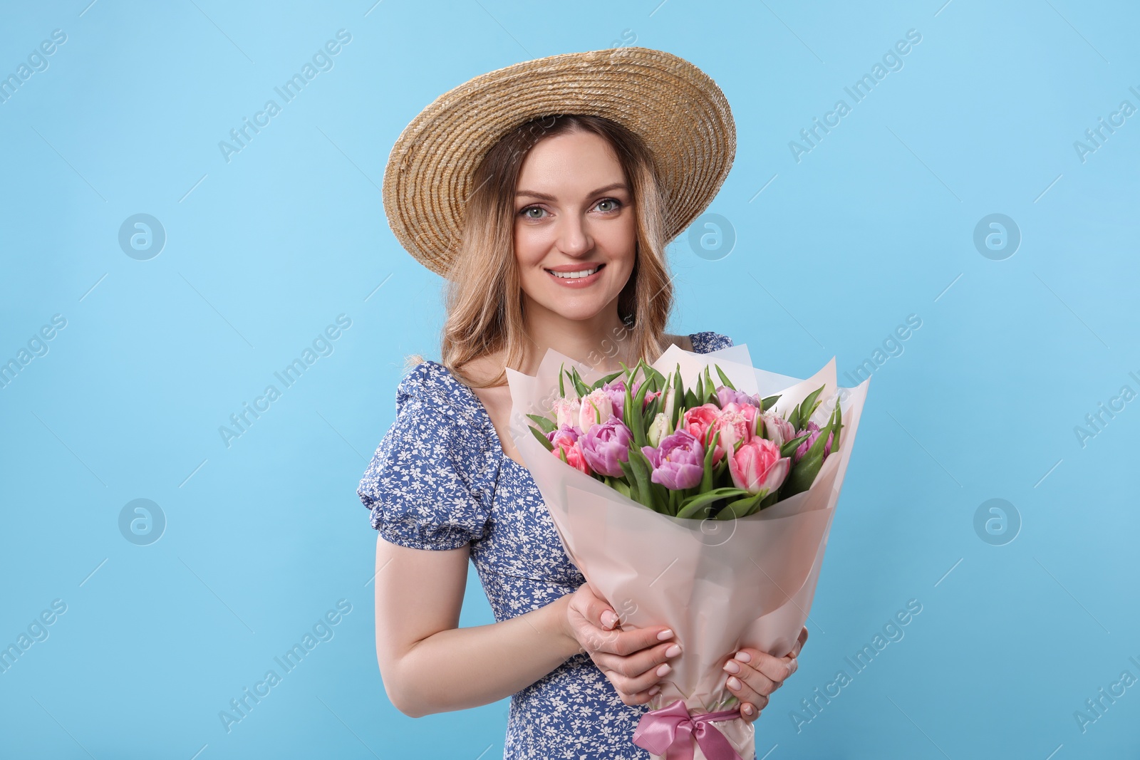 Photo of Happy young woman in straw hat holding bouquet of beautiful tulips on light blue background