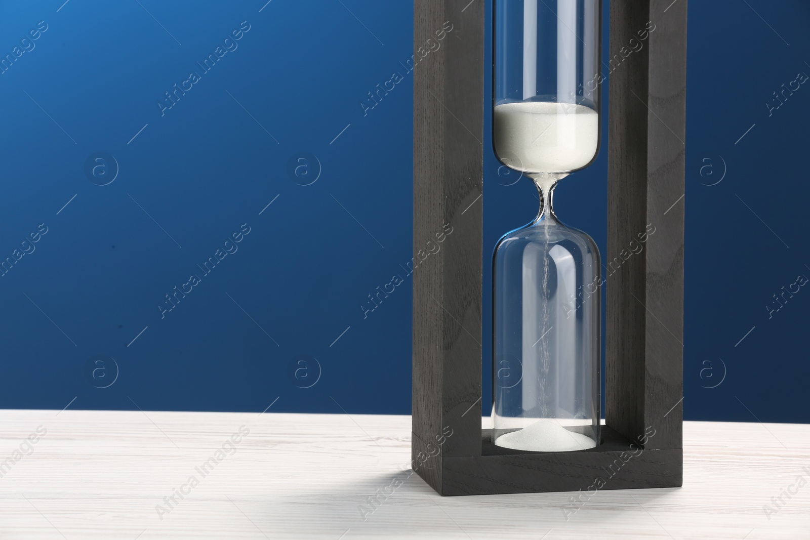 Photo of Hourglass with light green flowing sand on white table against blue background. Space for text