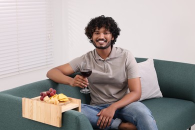 Photo of Happy man holding glass of wine at home. Grapes and snacks on sofa armrest wooden table