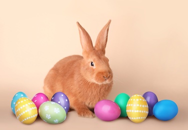 Cute bunny and wicker basket with bright Easter eggs on beige background