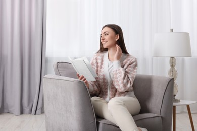 Woman reading book on armchair near window at home