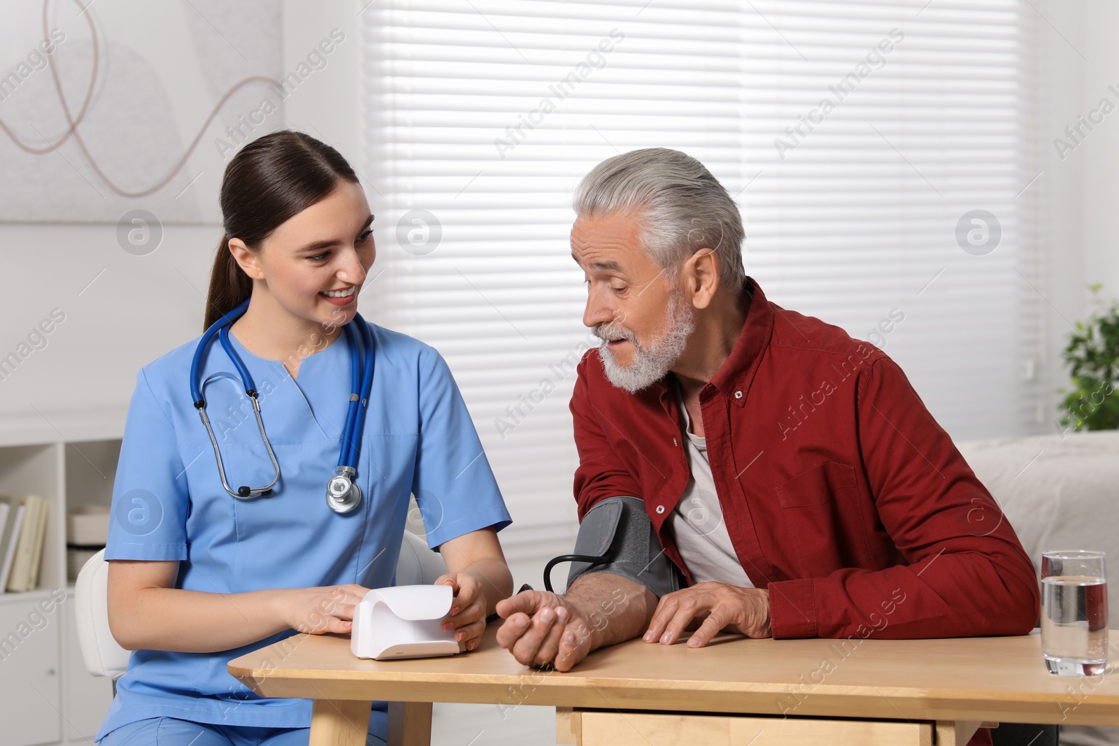 Photo of Young healthcare worker measuring senior man's blood pressure at wooden table indoors