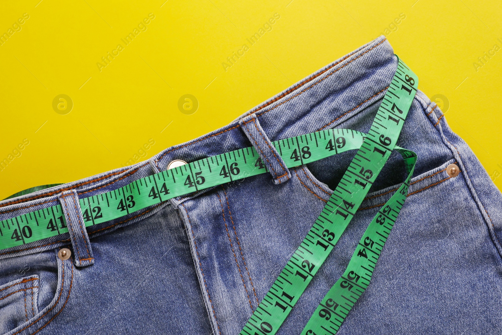 Photo of Jeans with measuring tape on yellow background, top view. Weight loss concept