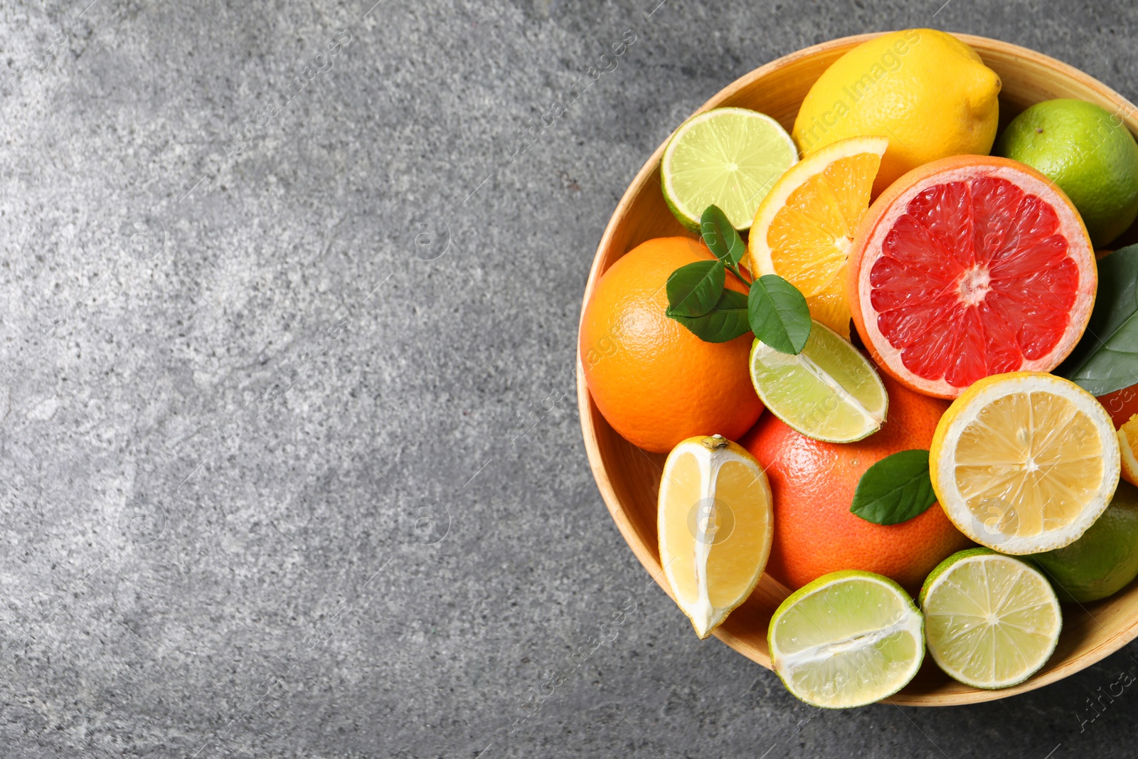 Photo of Different fresh citrus fruits and leaves in bowl on grey textured table, top view. Space for text