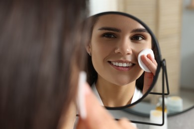 Photo of Young woman cleaning her face with cotton pad near mirror indoors
