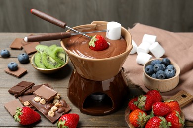 Photo of Fondue pot with melted chocolate, different fresh berries, kiwi, marshmallows and forks on wooden table