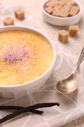 Photo of Delicious creme brulee in bowl, vanilla pods and spoon on table, closeup