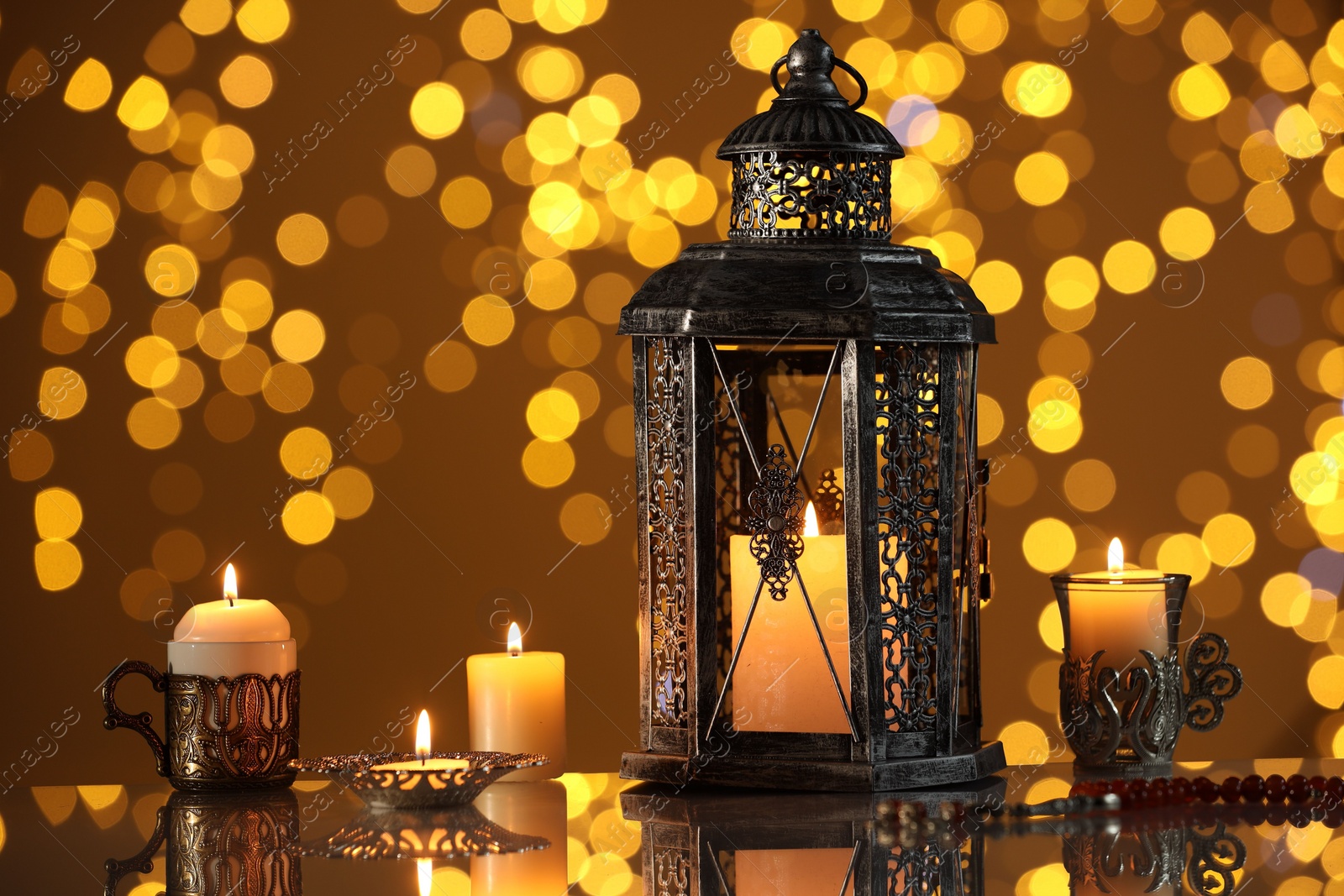 Photo of Arabic lantern and burning candles on mirror surface against blurred lights