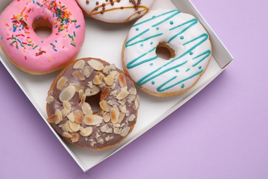 Photo of Box with different tasty glazed donuts on violet background, top view