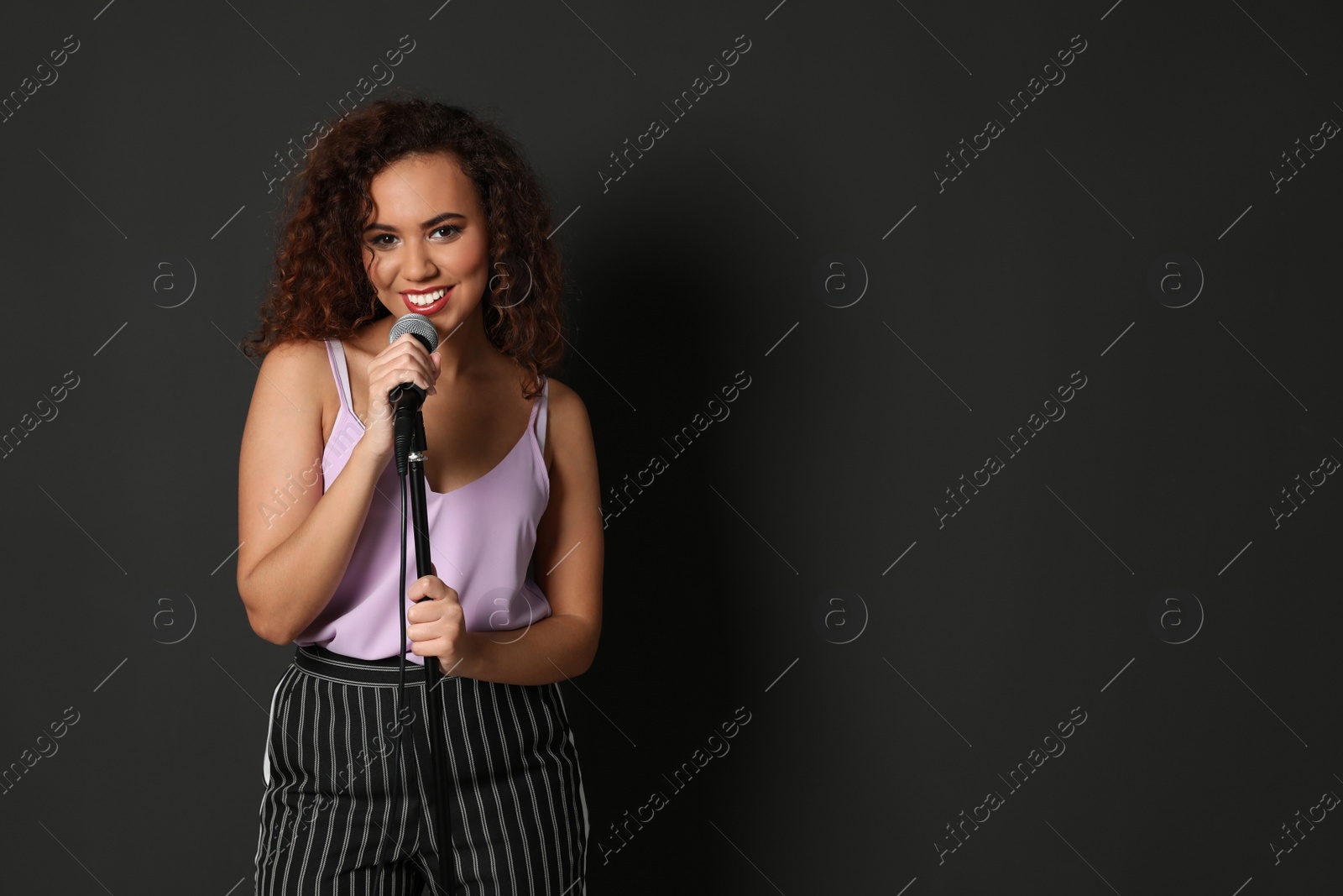 Photo of Curly African-American woman in stylish clothes posing with microphone on black background. Space for text