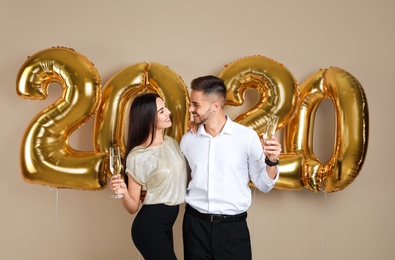 Photo of Happy young couple with glasses of champagne near golden 2020 balloons on beige background. New Year celebration