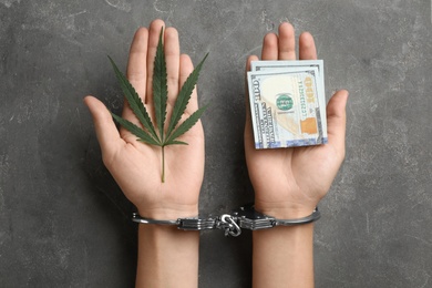 Photo of Woman in handcuffs holding hemp leaf and dollars on grey stone background, closeup