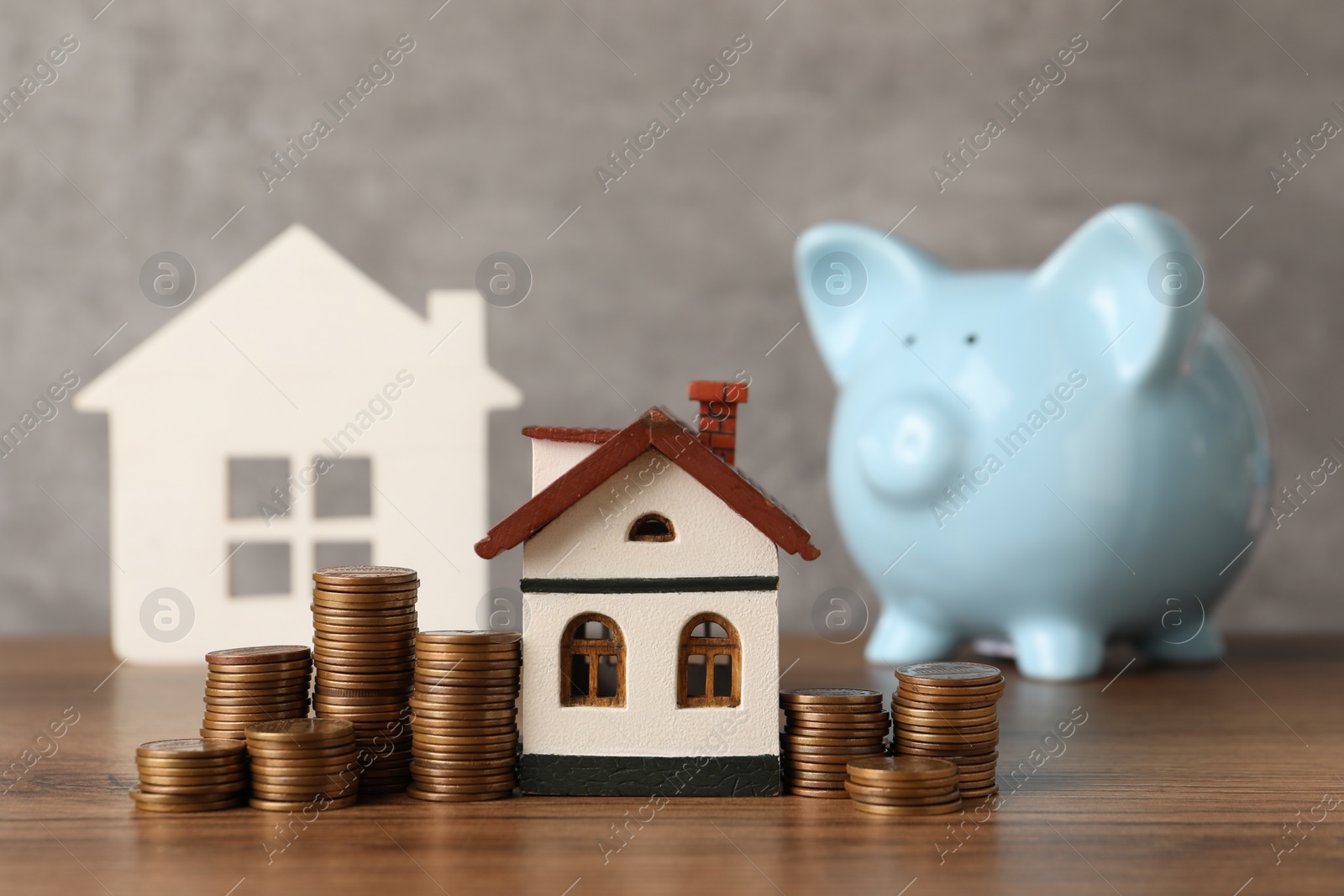 Photo of House models, piggy bank and stacked coins on wooden table against gray background, selective focus