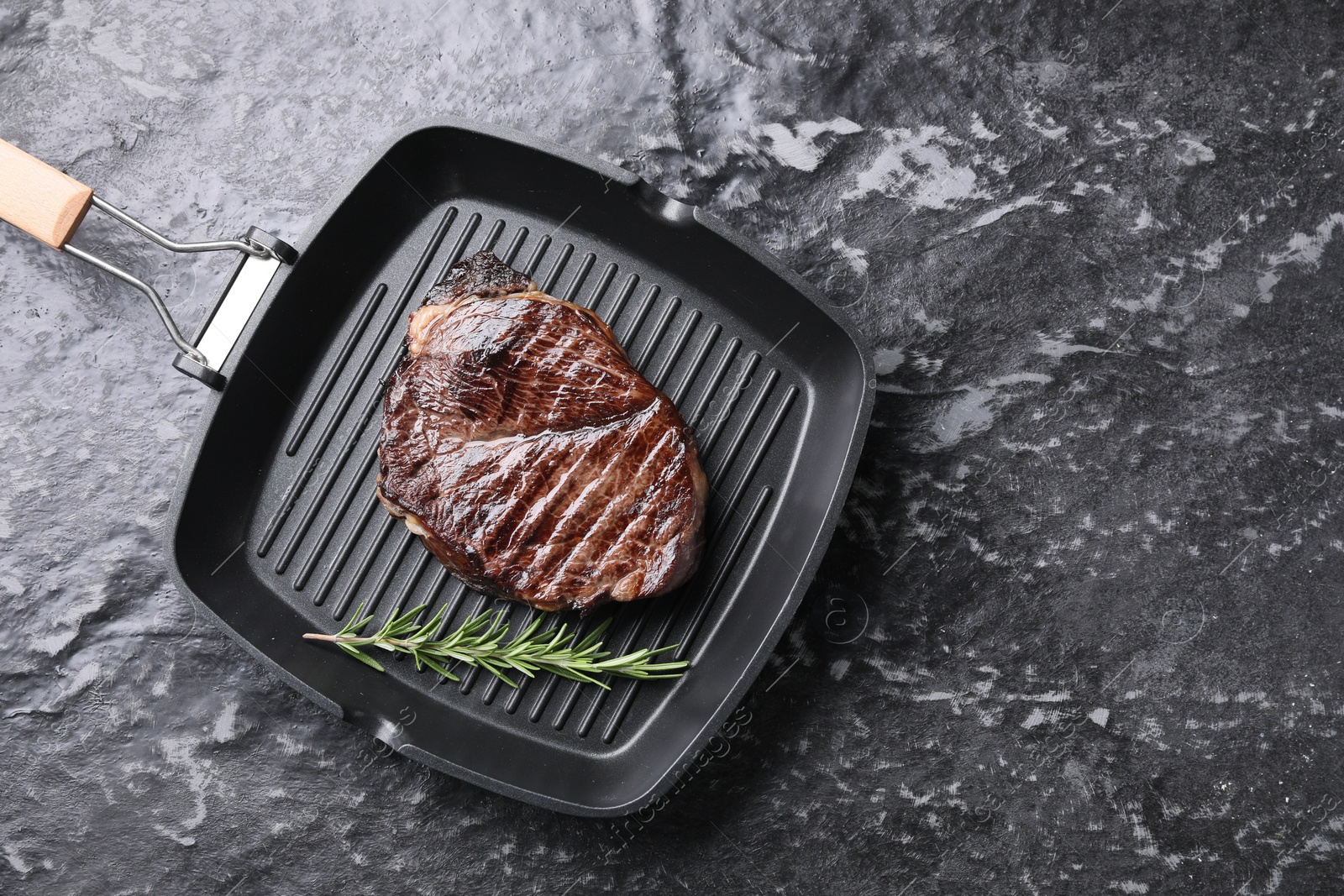 Photo of Grill pan with delicious fried beef meat and rosemary on grey textured table, top view. Space for text
