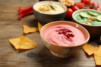 Different kinds of tasty hummus with nachos and ingredients on wooden table