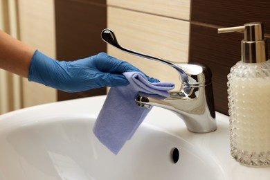 Photo of Woman in gloves cleaning faucet of bathroom sink with rag, closeup