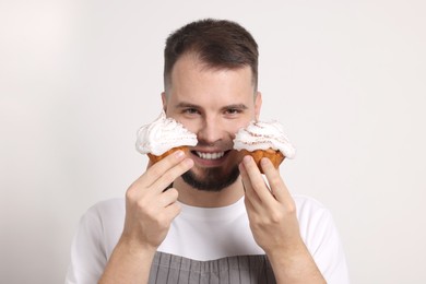 Photo of Happy professional confectioner in apron holding delicious cupcakes on light grey background