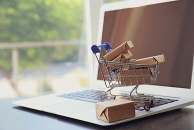 Photo of Internet shopping. Small cart with boxes and modern laptop on table indoors, space for text