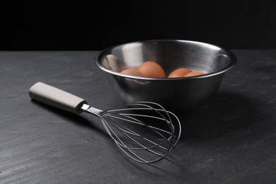 Metal whisk and raw eggs in bowl on dark grey table, closeup