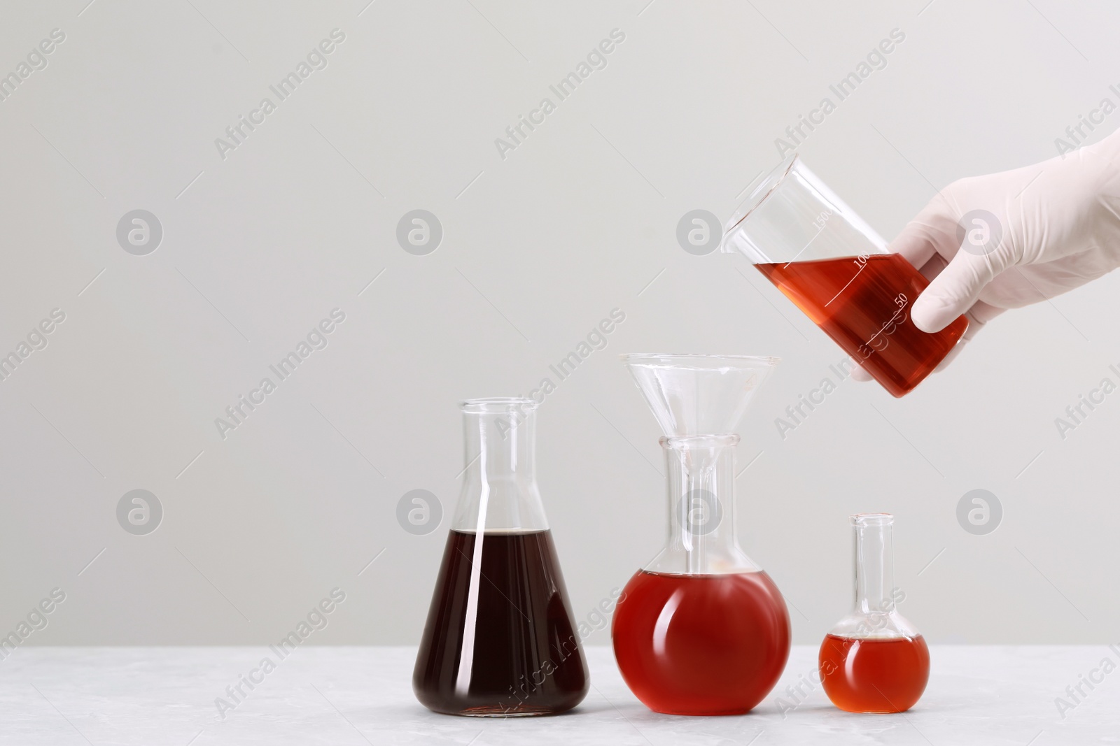 Photo of Scientist pouring liquid from beaker into round bottom flask on white table against light background, closeup. Space for text