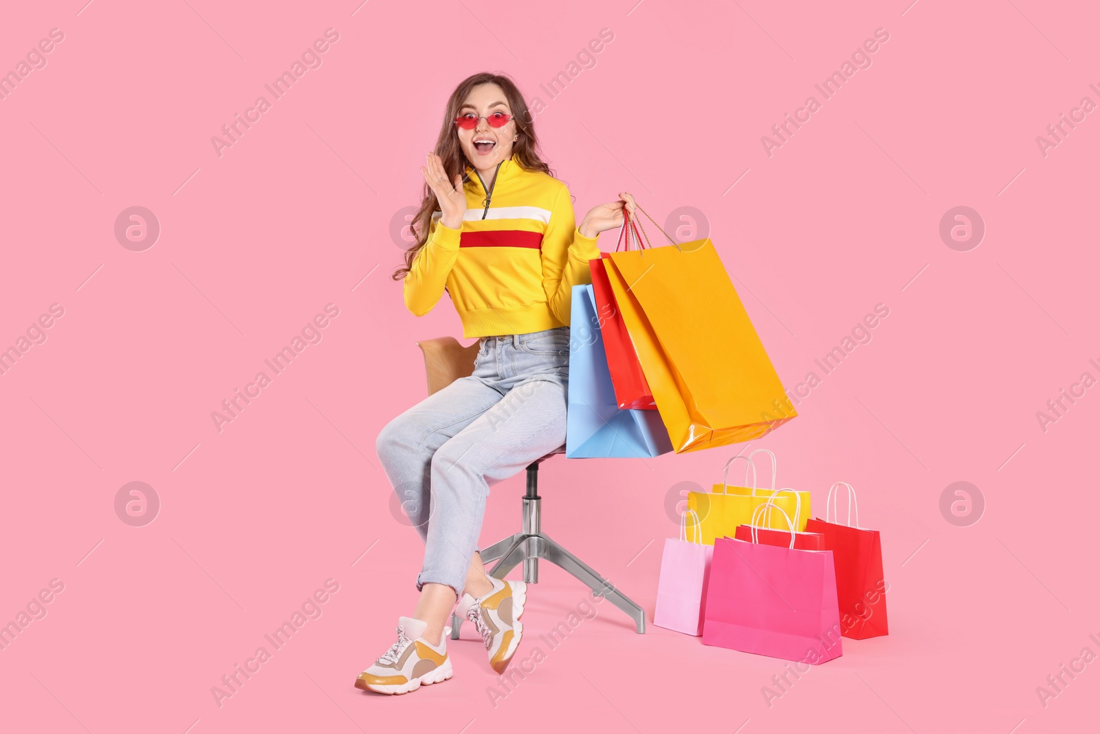 Photo of Emotional woman in stylish sunglasses holding many colorful shopping bags on armchair against pink background