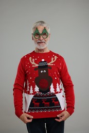 Photo of Senior man in funny glasses showing his Christmas sweater on grey background