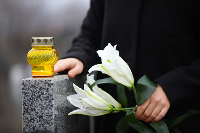 Woman holding white lilies near grey granite tombstone with candle outdoors, closeup. Funeral ceremony