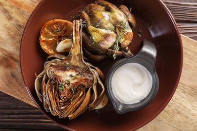 Photo of Tasty grilled artichoke and sauce in bowl on wooden table, top view