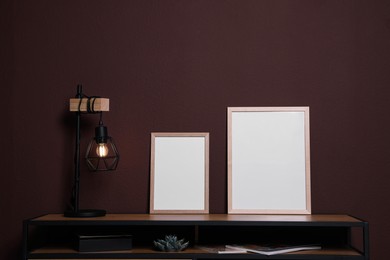 Photo of Empty frames and stylish lamp on wooden table near brown wall. Mockup for design