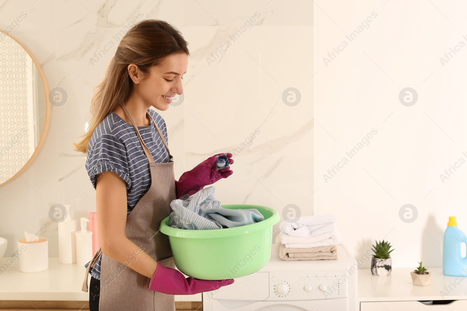 Photo of Woman holding dirty clothes and laundry detergent capsule in bathroom