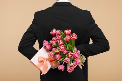 Photo of Man hiding beautiful bouquet and present on beige background, back view