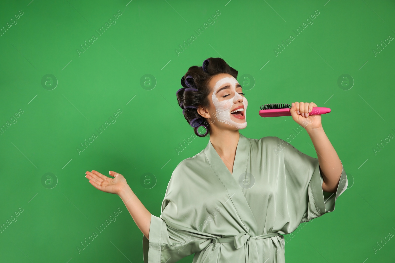 Photo of Happy young woman in silk bathrobe with hair curlers and facial mask singing into hairbrush on green background