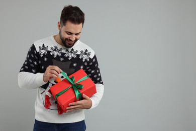 Young man in Christmas sweater opening gift on grey background. Space for text