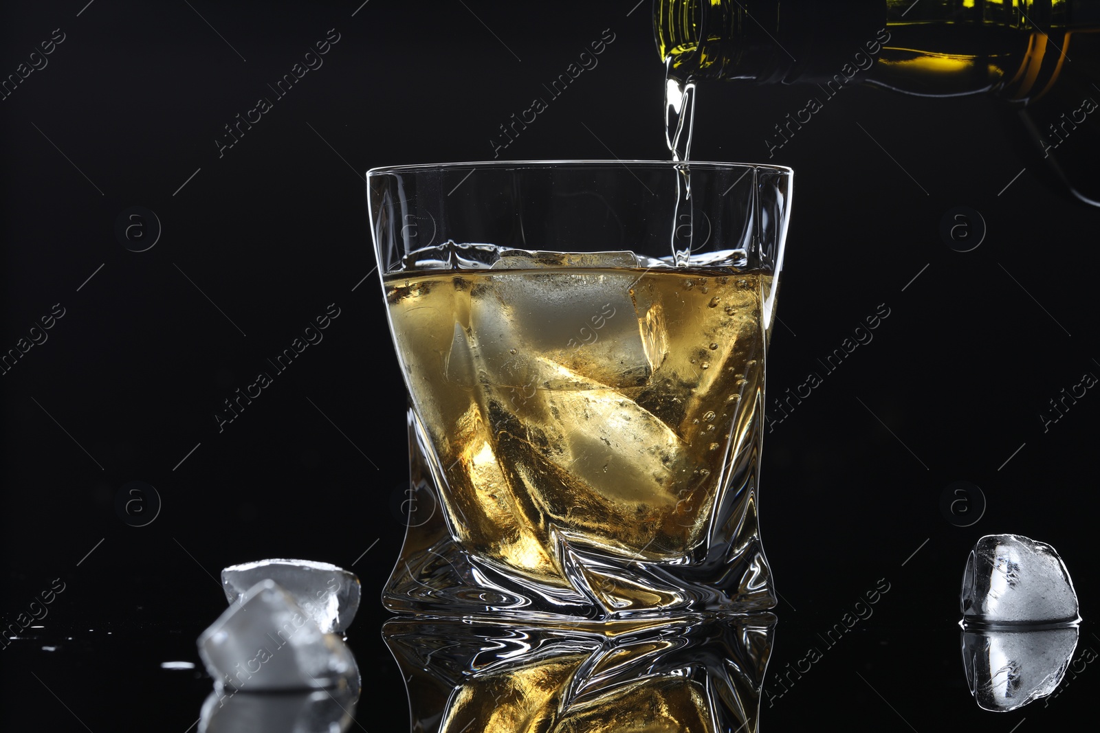 Photo of Pouring tasty whiskey from bottle into glass with ice at mirror table against black background, closeup