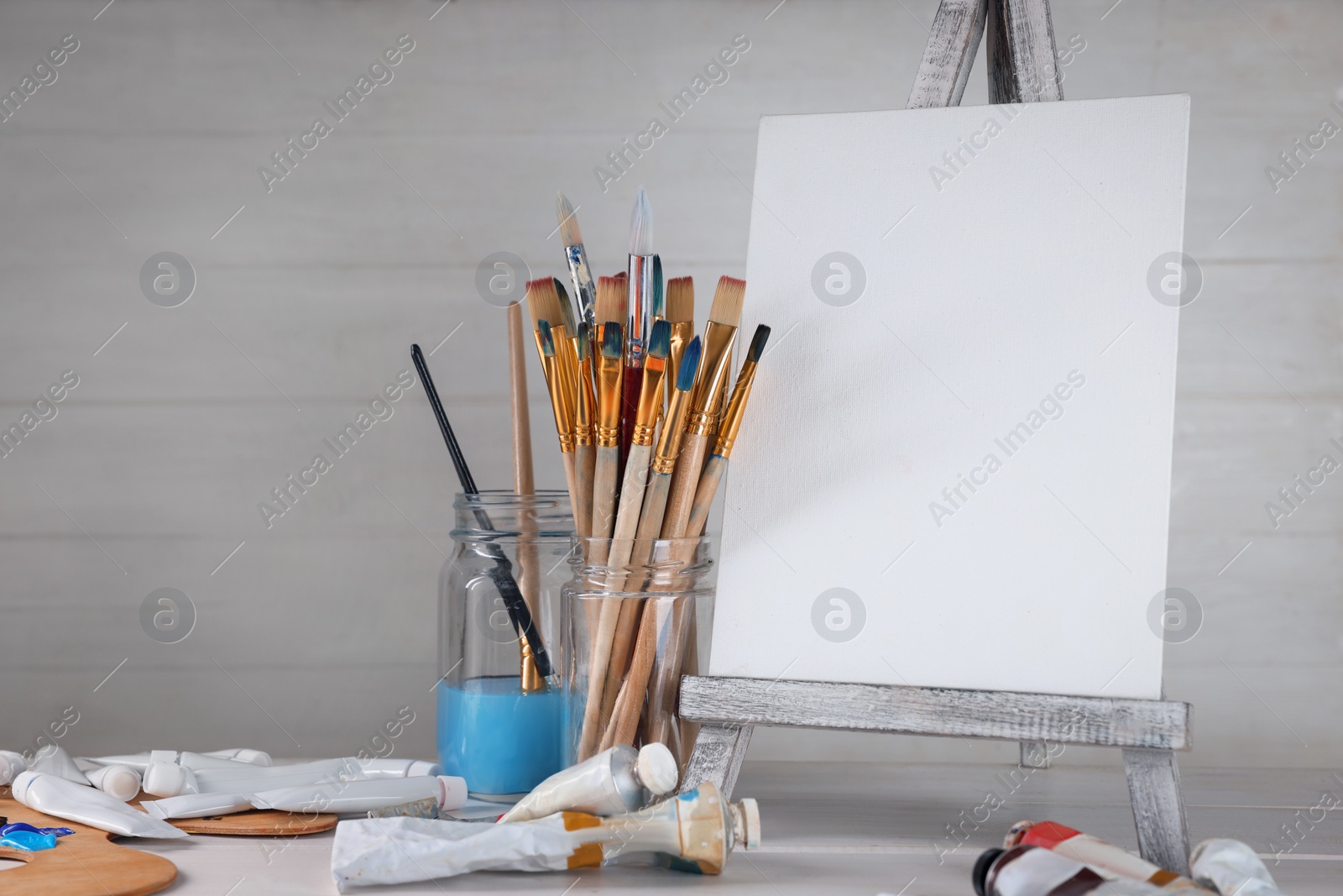 Photo of Many brushes, easel with blank canvas and paints on white table