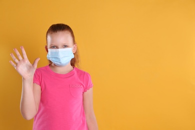 Photo of Little girl in protective mask showing hello gesture on yellow background, space for text. Keeping social distance during coronavirus pandemic