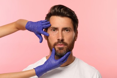 Doctor checking patient's face before cosmetic surgery operation on pink background