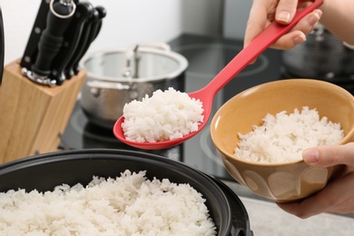 Photo of Woman putting boiled rice into bowl from cooker in kitchen, closeup