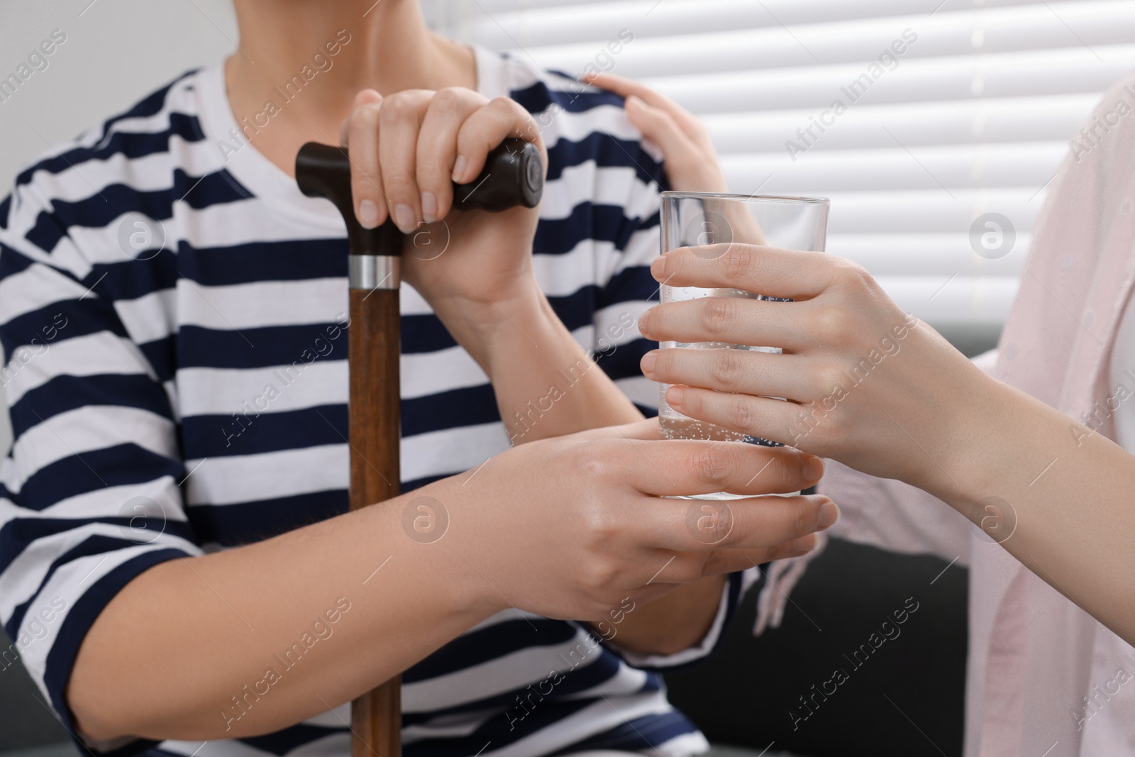 Photo of Caregiver giving water to senior woman with walking cane on sofa at home, closeup