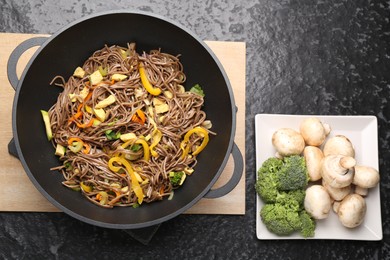 Photo of Stir-fry. Tasty noodles with vegetables, meat in wok and ingredients on dark textured table, flat lay