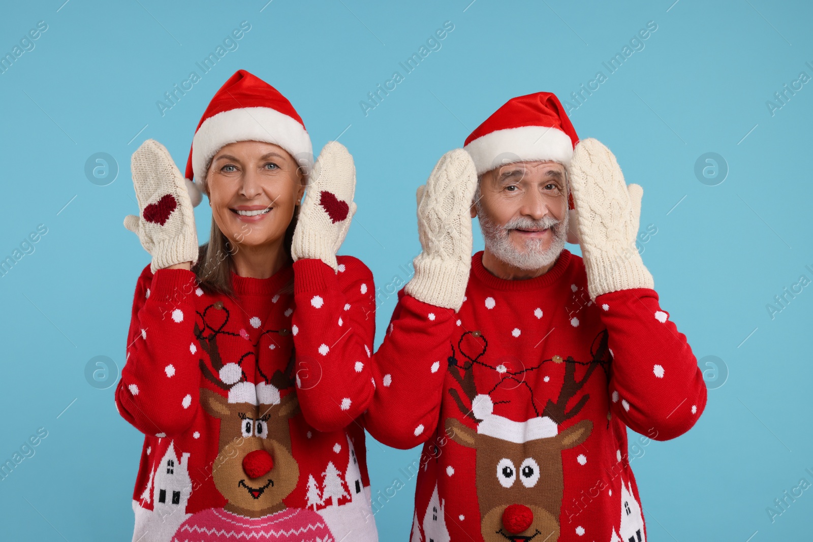 Photo of Happy senior couple in Christmas sweaters, Santa hats and knitted mittens on light blue background