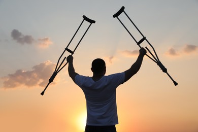 Photo of Man raising hands with underarm crutches up to sky outdoors at sunset, back view. Healing miracle