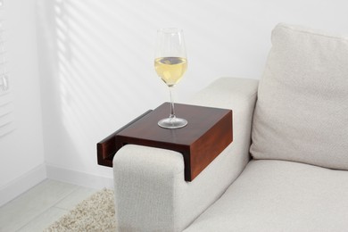 Photo of Glass of white wine on sofa with wooden armrest table in room. Interior element