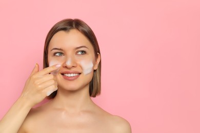 Photo of Young woman washing face with cleansing foam on pink background, space for text. Skin care cosmetic